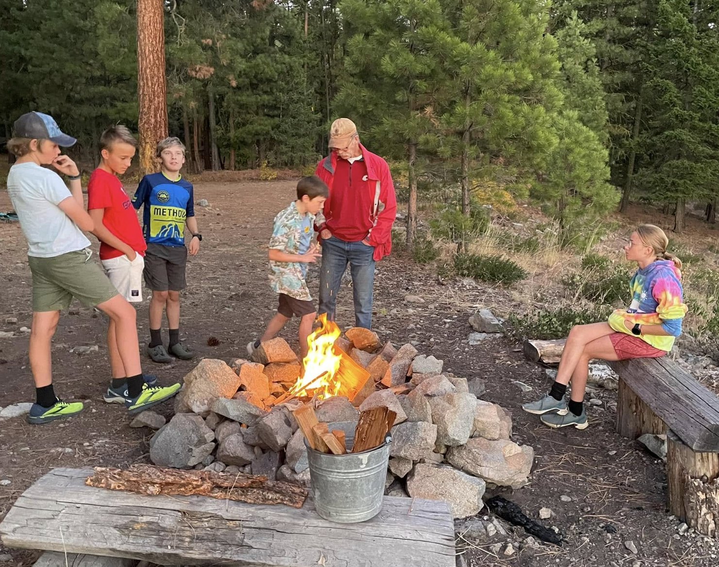 Youth gathered around a stone fire ring in a pine forest at dusk